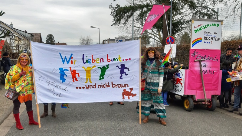 Zwei Narren im Kostüm halten ein Banner mit dem Schriftzug "Wir lieben bunt und nicht braun". Danaben der Aktionswagen von #EgelsbachIstMehr mit dem Spruch "Liebes Deutschalnd, kannst Du nicht mal nach den RECHTEN sehen?"