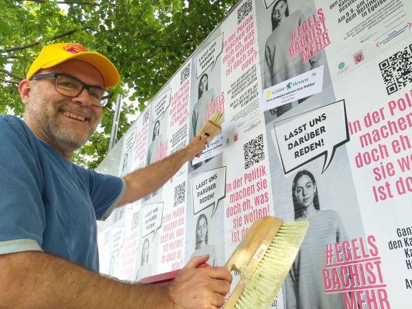 Ein in die Kamera lachern der Mann klebt die Aktioensplakate mehrfach auf eine Plakatwand